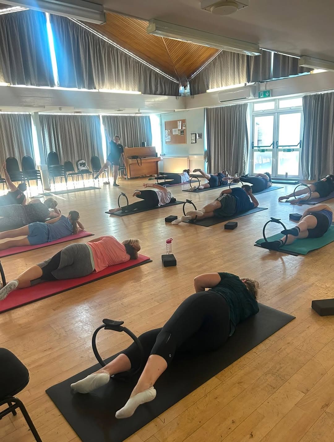 Mat Pilates class on mats with Pilates rings in a parquet-floored community hall