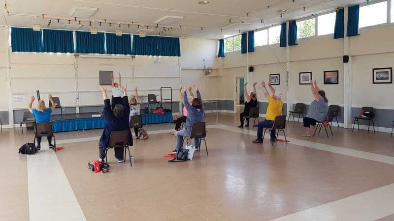 Chair based Pilates class, seated members with arms raised in a community hall